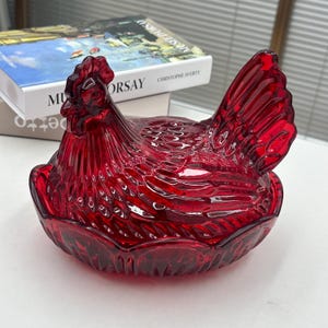 May include: A ruby red glass hen-shaped dish with a textured surface. The dish is in the shape of a hen, with a detailed comb, wattle, and tail feathers. The dish is likely used for decorative purposes or as a serving piece.