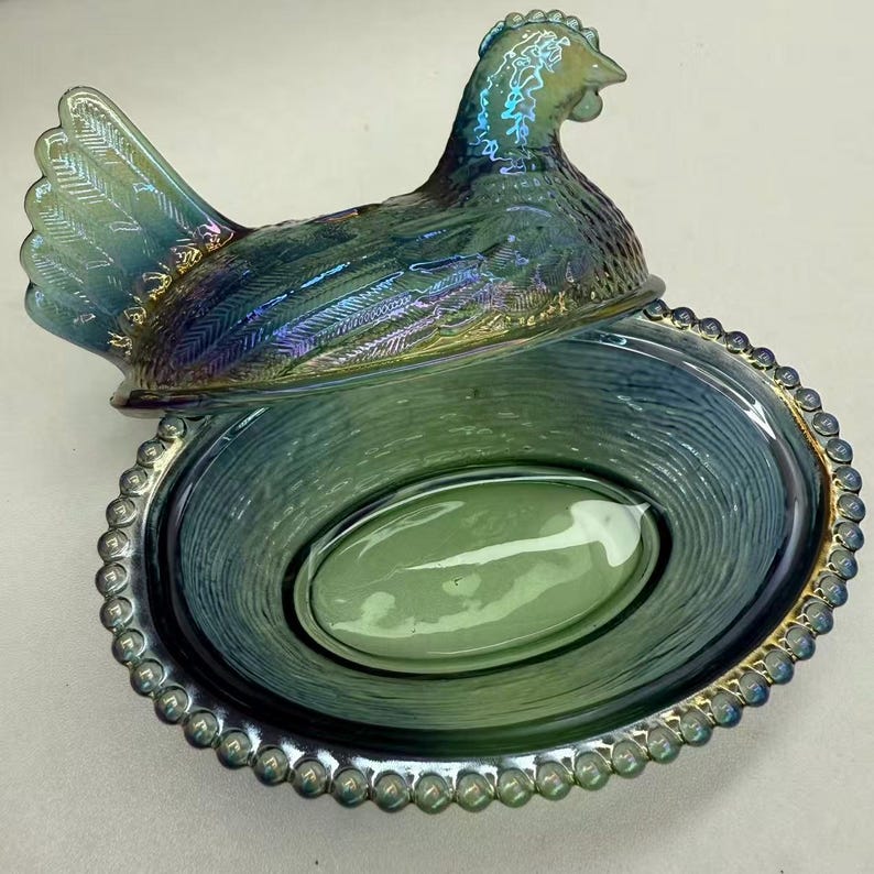 May include: Iridescent blue and green hen-shaped dish with a matching lid. The dish has an oval shape with a beaded rim. The hen lid features detailed feathering and a textured surface. The dish is designed to hold an egg.