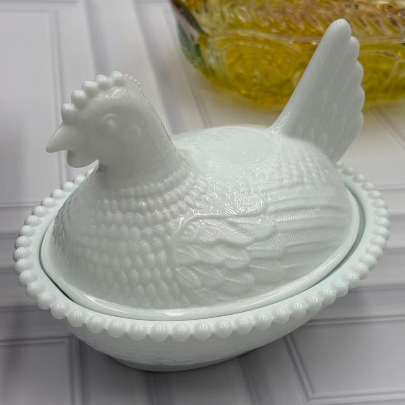 May include: A white milk glass hen on nest dish featuring a detailed molded chicken lid with textured feather patterns and a beaded rim base. The opaque white glass container is shaped like a nesting hen with an upright tail and a defined comb.