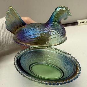 May include: Iridescent glass hen-shaped covered dish with a matching oval base. The hen is detailed with textured feathers and a comb, displaying shades of blue, green, and purple. The base has a beaded edge, creating a decorative serving piece.