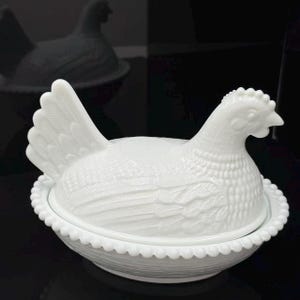 May include: A white milk glass hen on a nest dish. The lid is shaped like a sitting hen with detailed feather textures, and the base is a round bowl with a beaded rim and a woven basket pattern on the exterior, creating a classic lidded container.