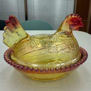 May include: A decorative, iridescent glass hen-shaped dish with a lid. The dish is primarily yellow with red and purple accents, featuring a textured surface and a beaded rim. The hen's head and tail are detailed with vibrant colors.