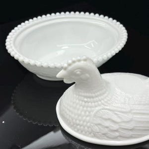 May include: A white milk glass hen on nest dish featuring a detailed chicken-shaped lid with textured feather patterns and a matching oval base with a beaded rim edge. The lid is positioned in front of the open, empty, smooth-bottomed bowl.