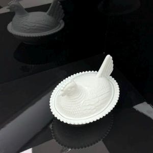 May include: A white milk glass hen on a nest dish featuring a detailed molded chicken lid with a textured feather pattern and a beaded rim edge. The oval-shaped container is designed with a nesting base and a lid shaped like a sitting hen.