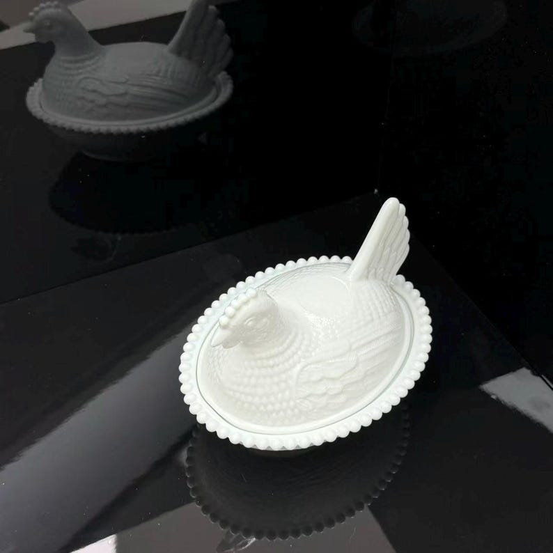 May include: A white milk glass hen on a nest dish featuring a detailed molded chicken lid with a textured feather pattern and a beaded rim edge. The oval-shaped container is designed with a nesting base and a lid shaped like a sitting hen.