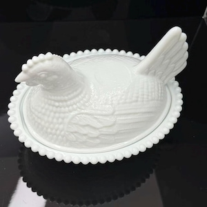 May include: A white milk glass hen on a nest dish featuring a detailed molded chicken figure with textured feathers and a comb, resting on an oval base with a decorative beaded edge. The lid is shaped like a sitting hen with a raised tail.