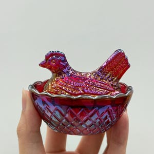 May include: A small, iridescent red glass hen-on-nest dish. The lid is shaped like a sitting hen with detailed feather textures, while the base features a diamond-patterned, cross-hatched design. The glass has a metallic, multi-colored sheen.