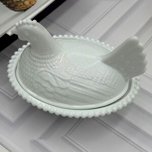 May include: A white milk glass hen on a nest dish featuring a detailed molded chicken lid with textured feather patterns and a beaded rim base. The opaque glass container is shaped like a sitting hen with a raised head and tail, resting on an oval dish.