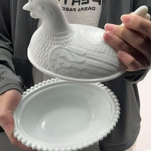 May include: A white milk glass hen on nest dish featuring a detailed molded chicken lid with textured feather patterns and a matching oval base with a beaded rim edge. The lid is lifted to reveal the smooth, hollow interior of the serving bowl.