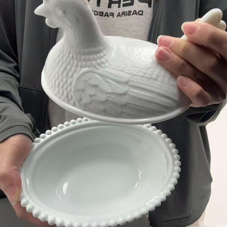 May include: A white milk glass hen on nest dish featuring a detailed molded chicken lid with textured feather patterns and a matching oval base with a beaded rim edge. The lid is lifted to reveal the smooth, hollow interior of the serving bowl.