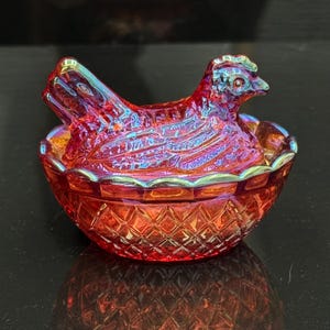 May include: A red iridescent glass hen on a nest dish featuring a pressed diamond pattern on the base and detailed feather textures on the lid. The glass displays a shimmering rainbow finish across the surface of the chicken-shaped cover.