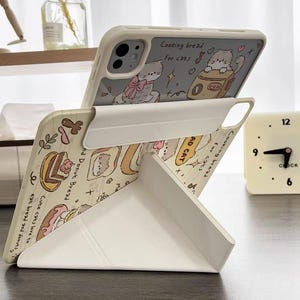 Puede incluir: Una funda de teléfono blanca con soporte, con un diseño de gato de dibujos animados y el texto "Cooking bread for cats". La funda está apoyada en un escritorio, con un pequeño reloj al fondo.