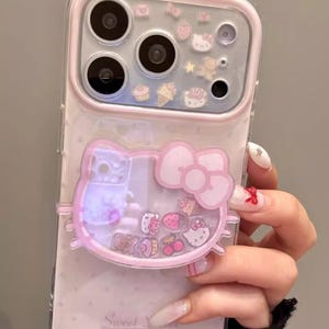 Puede incluir: Una funda de teléfono transparente y rosa con un diseño de Hello Kitty. La funda tiene un borde rosa y una parte trasera transparente con abalorios flotantes. El diseño incluye una cara de Hello Kitty, un lazo y otros pequeños abalorios.