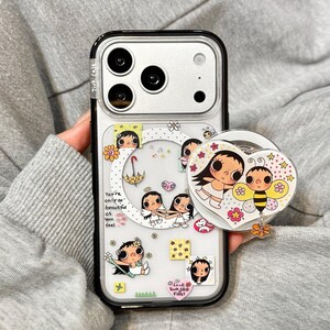 Può includere: Custodia trasparente per telefono con bordo nero, con illustrazioni di cartoni animati di ragazze, fiori e un'ape. La custodia include il testo "You're only as beautiful as you feel" e un supporto per telefono a forma di cuore.