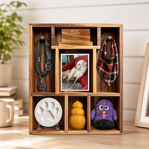 May include: A wooden pet memorial shadow box featuring a black collar with a circular tag, a plaid bandana, a photograph of a dog on a red sofa, a white paw print casting, a yellow rubber toy, and a purple plush toy with the name Boby engraved.