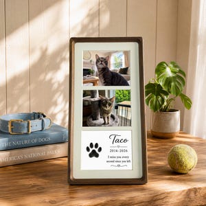 May include: A vertical wooden picture frame containing two photographs of a cat and a white memorial card. The card features a black paw print, the name Taco, the years 2014-2026, and the text I miss you every second since you left.