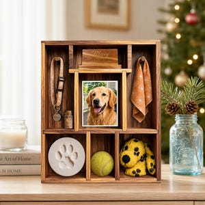 Op de afbeelding: Een houten vitrinekast met meerdere vakken. Het bevat een halsband voor huisdieren met een penning, een klein glazen potje, een foto van een golden retriever, een pootafdruk, een tennisbal en een geel pluche hondenspeeltje.
