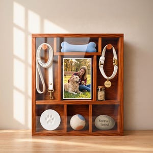 May include: A wooden shadow box display case featuring a white leash, a light blue bone-shaped toy, a white collar with a gold tag reading Buddy, a small glass jar, a white paw print casting, a ball, and a stone engraved with forever loved.
