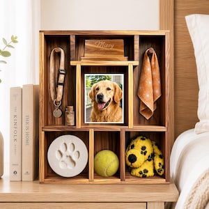May include: A wooden pet memorial shadow box with multiple compartments. It contains a photo of a golden retriever, a dog collar, a small glass jar, a paw print casting, a tennis ball, a plush dog toy, and a wooden plaque engraved with Missy.