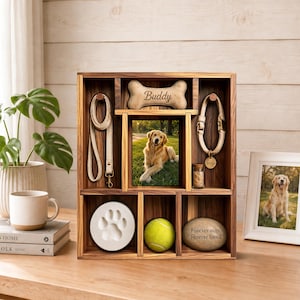 May include: A wooden shadow box display case featuring a central photo of a golden retriever, a beige leash, a collar with a tag reading Buddy, a bone-shaped plaque labeled Buddy, a white paw print casting, a tennis ball, and a memorial stone.