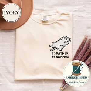 May include: Cream-colored t-shirt with an embroidered design of a gray cat lying down. The text "I'D RATHER BE NAPPING" is below the cat. The shirt has a crew neck and short sleeves. The shirt is XL size.