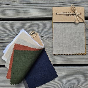 May include: A fabric swatch sample set featuring multiple colours including white, beige, rust orange, olive green, and navy blue. The swatches are bound with a brown cardboard header labelled NEXT TO LINEN and tied with a thin jute twine bow.