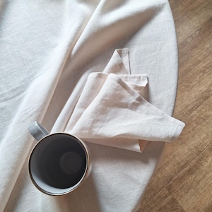 May include: A light beige linen tablecloth with a matching folded napkin placed on top. An empty ceramic mug with a grey exterior and a light-colored interior sits on the fabric. The textile features a visible woven texture and soft folds.