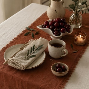 Peut inclure: Un chemin de table en lin couleur rouille aux bords effilochés posé sur une nappe blanche. Le chemin est décoré d'un bol en céramique rempli de cerises rouge foncé, d'une tasse mouchetée, d'une assiette avec une serviette en lin, une fourchette dorée et une bougie.
