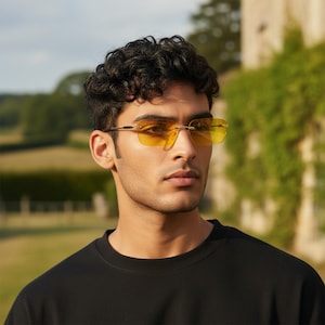 May include: A person wearing rimless sunglasses with yellow tinted lenses and gold-coloured frames. The person is wearing a black crew neck top. The background features a green landscape and a building.