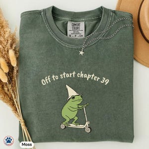 Puede incluir: Camiseta verde musgo con el texto "Off to start chapter 39" y una rana de dibujos animados en un patinete. La rana lleva un sombrero de mago y gafas de sol. Un collar plateado con un colgante de estrella está sobre la camiseta.
