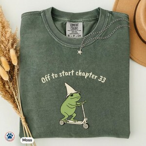 May include: Moss green t-shirt with the text "Off to start chapter 33" and a cartoon frog riding a scooter. The frog wears a wizard hat. A silver chain with a star pendant is draped on the shirt.