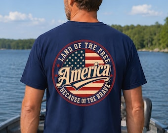 Peut inclure: Un t-shirt bleu marine à manches courtes avec un motif circulaire au dos. Le design comprend un drapeau américain stylisé avec le texte "Land of the Free" et "Because of the Brave" entourant le mot "America" dans une police cursive.