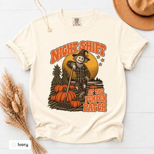 May include: An ivory short-sleeve t-shirt featuring a graphic print of a scarecrow in a pumpkin patch with a rake. The text reads "NIGHT SHIFT AT THE PUMPKIN PATCH" in orange letters. The design includes a large yellow moon and corn stalks.