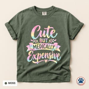 Könnte beinhalten: Ein moosgrünes Kurzarm-T-Shirt mit dem Text "Cute BUT MEDICALLY Expensive" in einem mehrfarbigen Pastell-Tie-Dye-Druck. Das Design enthält dekorative Schnörkel und ein kleines Herz unter dem Text auf der Vorderseite des T-Shirts.