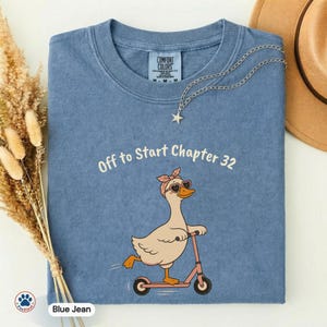 May include: Blue jean colored t-shirt with the text "Off to Start Chapter 32" and a cartoon goose wearing heart-shaped sunglasses and a pink headband, riding a pink scooter. A silver necklace with a star pendant is draped on the shirt.