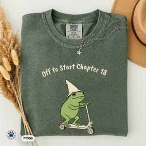 Può includere: T-shirt verde muschio con la scritta "Off to Start Chapter 18" e un ranocchio dei cartoni animati su uno scooter, con un cappello da mago. Una collana d'argento con un ciondolo a stella è drappeggiata sulla maglietta.