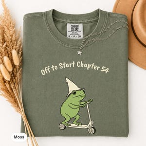 May include: A moss green crew neck t-shirt featuring a graphic of a green frog wearing a cream-colored pointed hat and riding a white scooter. The text above the frog reads "Off to Start Chapter 54" in a cream-colored font. The shirt has a Comfort Colors label.