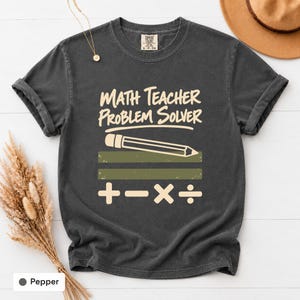 以下が含まれることがあります： クリーム色の「MATH TEACHER PROBLEM SOLVER」という文字がプリントされたチャコールグレーの半袖Tシャツ。文字の下には鉛筆のグラフィック、2本のオリーブグリーンの水平バー、そして足し算、引き算、掛け算、割り算の数学記号が描かれています。