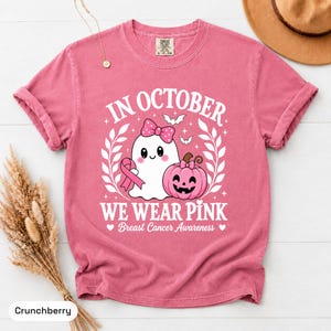 May include: A pink short-sleeve t-shirt featuring a graphic of a white ghost with a pink bow and a pink pumpkin. The text reads: IN OCTOBER WE WEAR PINK Breast Cancer Awareness. A pink ribbon is included in the design along with small bats.