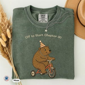May include: A moss green crewneck t-shirt featuring a brown bear wearing a striped party hat and riding a red tricycle. The text "Off to Start Chapter 30" is printed above the bear. The shirt has a Comfort Colors label at the inner neckline.