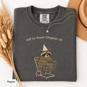 May include: A charcoal gray crewneck t-shirt featuring a graphic of a raccoon wearing a party hat inside a shopping cart. The text "Off to Start Chapter 31" is printed above the raccoon. The shirt has a Comfort Colors brand label at the neck.