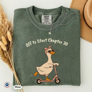 May include: Moss green t-shirt with the text "Off to Start Chapter 30" and a cartoon goose wearing sunglasses and a pink headband, riding a pink scooter. A silver necklace with a star pendant is draped on the shirt.