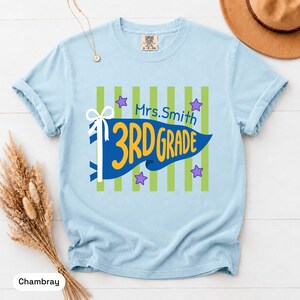 May include: A light blue t-shirt featuring a graphic with vertical lime green stripes, a blue pennant flag, and the yellow text 3RD GRADE. Above the flag, the text Mrs. Smith is printed in blue, accompanied by small purple stars and a white bow.