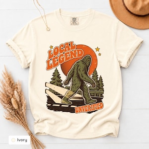 May include: An ivory short-sleeve t-shirt featuring a graphic of a green Bigfoot walking across a road. The text reads "LOCAL LEGEND" in orange above the creature and "UNVERIFIED" in orange below, set against a large orange sun and pine trees.