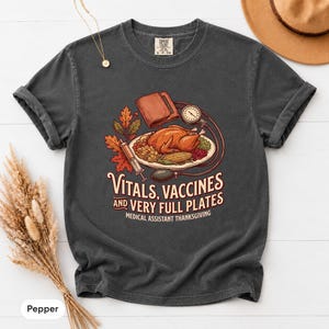 Puede incluir: Una camiseta gris carbón de manga corta con un gráfico de un pavo asado en un plato, una jeringuilla y un tensiómetro. El texto dice: VITALS, VACCINES AND VERY FULL PLATES MEDICAL ASSISTANT THANKSGIVING, rodeado de hojas de otoño.