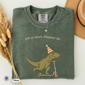 May include: A moss green crew neck t-shirt featuring a graphic of a dinosaur wearing a striped party hat and riding a scooter. The text "Off to Start Chapter 30" is printed above the dinosaur. The shirt has a vintage wash and a tag reading Comfort Colors.
