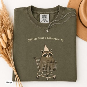 May include: An olive green t-shirt featuring a graphic of a raccoon wearing a party hat and sitting in a shopping cart. The text above the raccoon reads "Off to Start Chapter 18". The shirt has a Comfort Colors brand label at the inner neckline.