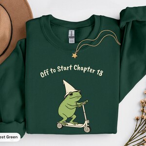 May include: A forest green long-sleeve sweatshirt featuring a graphic of a green frog wearing a cream-colored pointed hat and riding a white scooter. Above the frog, the text reads "Off to Start Chapter 18" in a cream-colored sans-serif font.