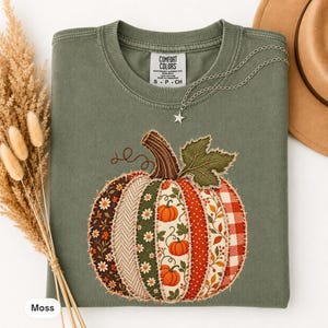 May include: An olive green crew neck t-shirt featuring a large pumpkin appliqué made of various fabric patches including floral, plaid, and polka dot patterns. The shirt has a Comfort Colors label at the neck and a silver star pendant necklace.