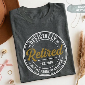 Op de afbeelding: Donkergrijs gemêleerd T-shirt met een cirkelvormige afbeelding met de tekst "Officially Retired" in witte en gouden letters. De tekst "EST. 2026" en "NOT MY PROBLEM ANYMORE" zijn ook inbegrepen. Het shirt heeft een ronde hals.